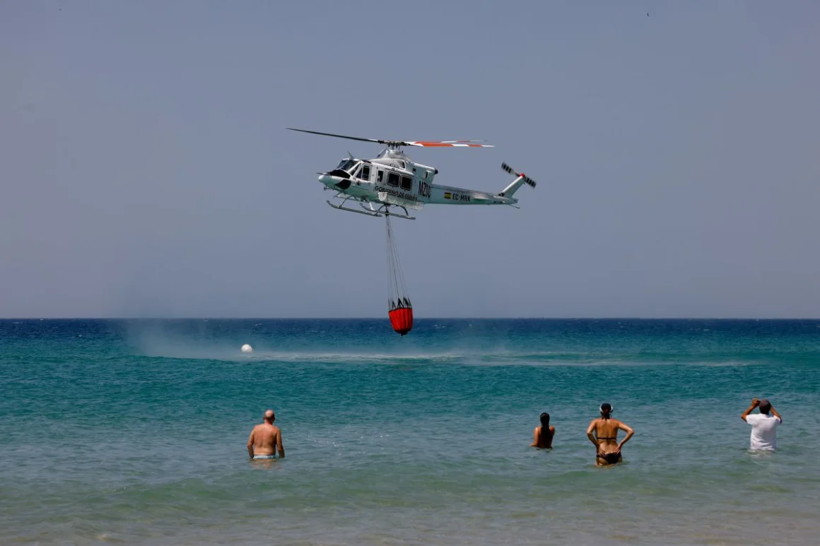 La gente observa cómo un helicóptero extrae agua en la playa de Atlanterra el 12 de agosto de 2025, un día después de que muchas personas, incluyendo residentes y turistas, fueran evacuadas de Tarifa debido a los incendios forestales. Marcelo del Pozo/Reuters