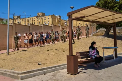 Una residente de Ceuta se sienta en una estación de autobuses mientras soldados españoles escoltan a migrantes que cruzaron a España hacia la frontera con Marruecos, para su regreso al país vecino desde el enclave español de Ceuta, el domingo 2 de agosto de 2026. Bernat Armangue/AP